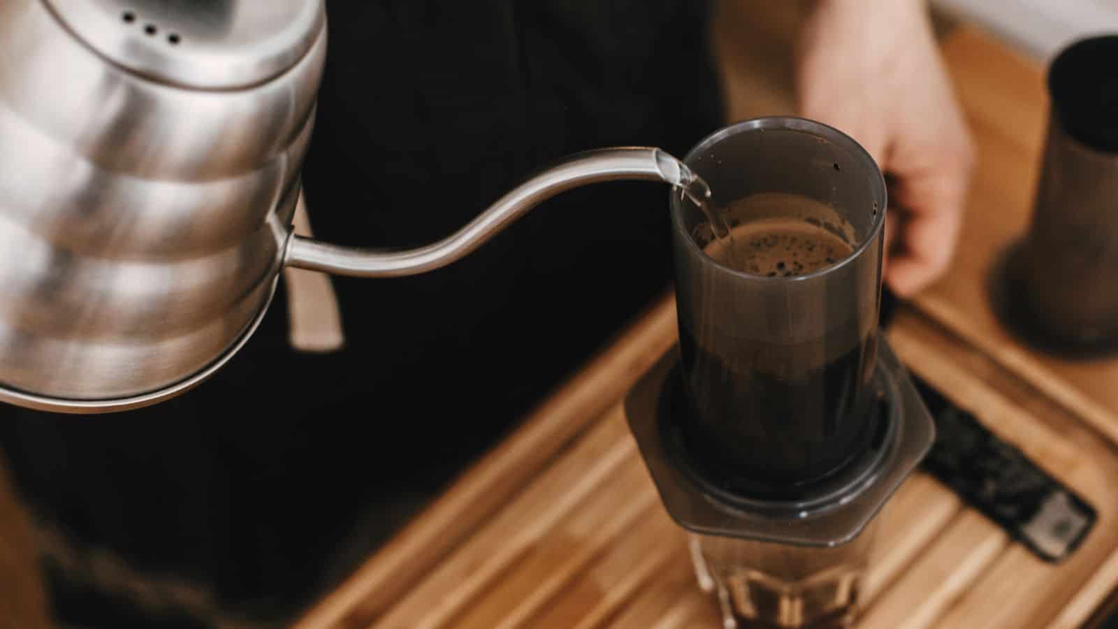 A person pours hot water from a gooseneck kettle into an AeroPress coffee maker on a wooden countertop. The AeroPress is partially filled with coffee grounds, and a dark liquid is visible inside.