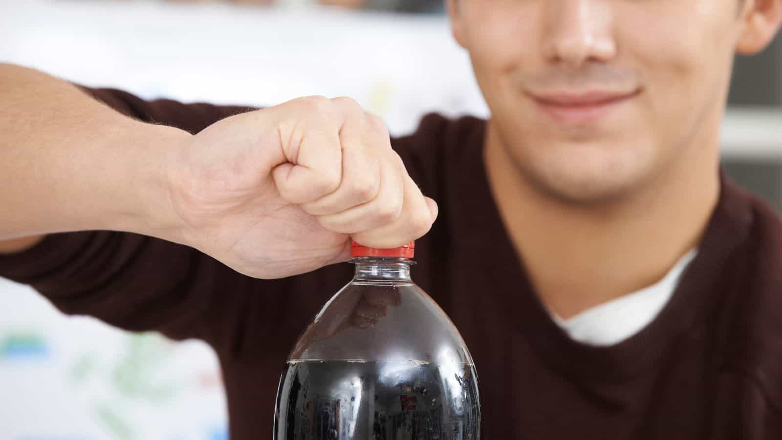 A person in a brown sweater is opening a plastic bottle with a red cap. The bottle appears to contain a dark, carbonated beverage. The person's face is partially visible, with a slight smile. Background details are blurred.