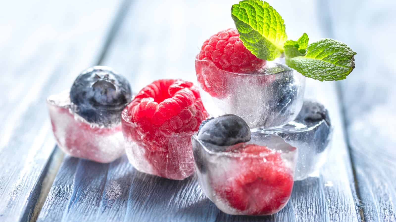 Several ice cubes containing raspberries and blueberries are stacked on a light blue wooden surface. A sprig of green mint leaves is placed on top of the ice cubes, adding a touch of color.
