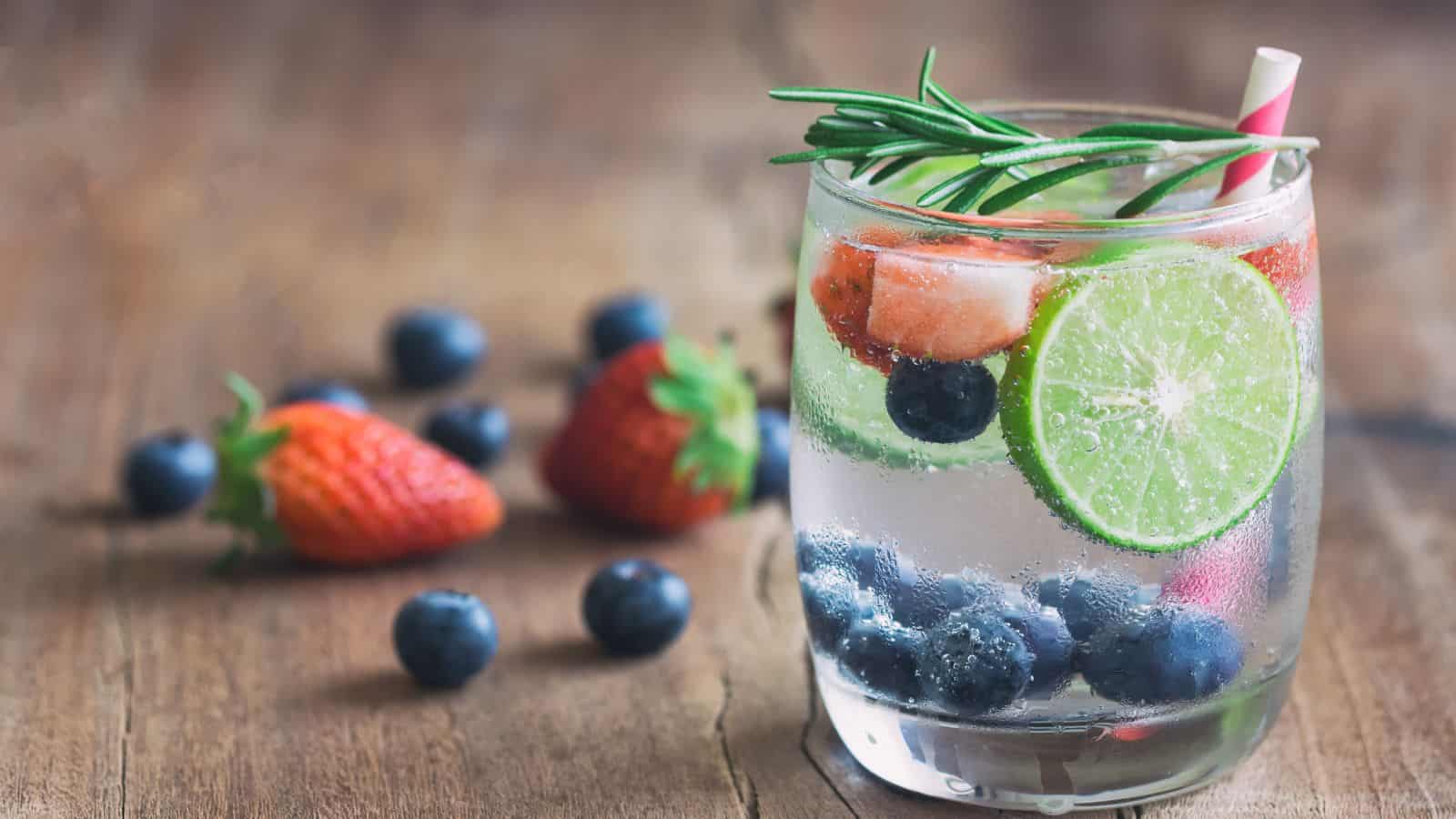 A glass of infused water on a wooden surface, containing blueberries, a lime slice, strawberries, and a sprig of rosemary. A striped straw is in the glass. Additional blueberries and strawberries are scattered in the background.