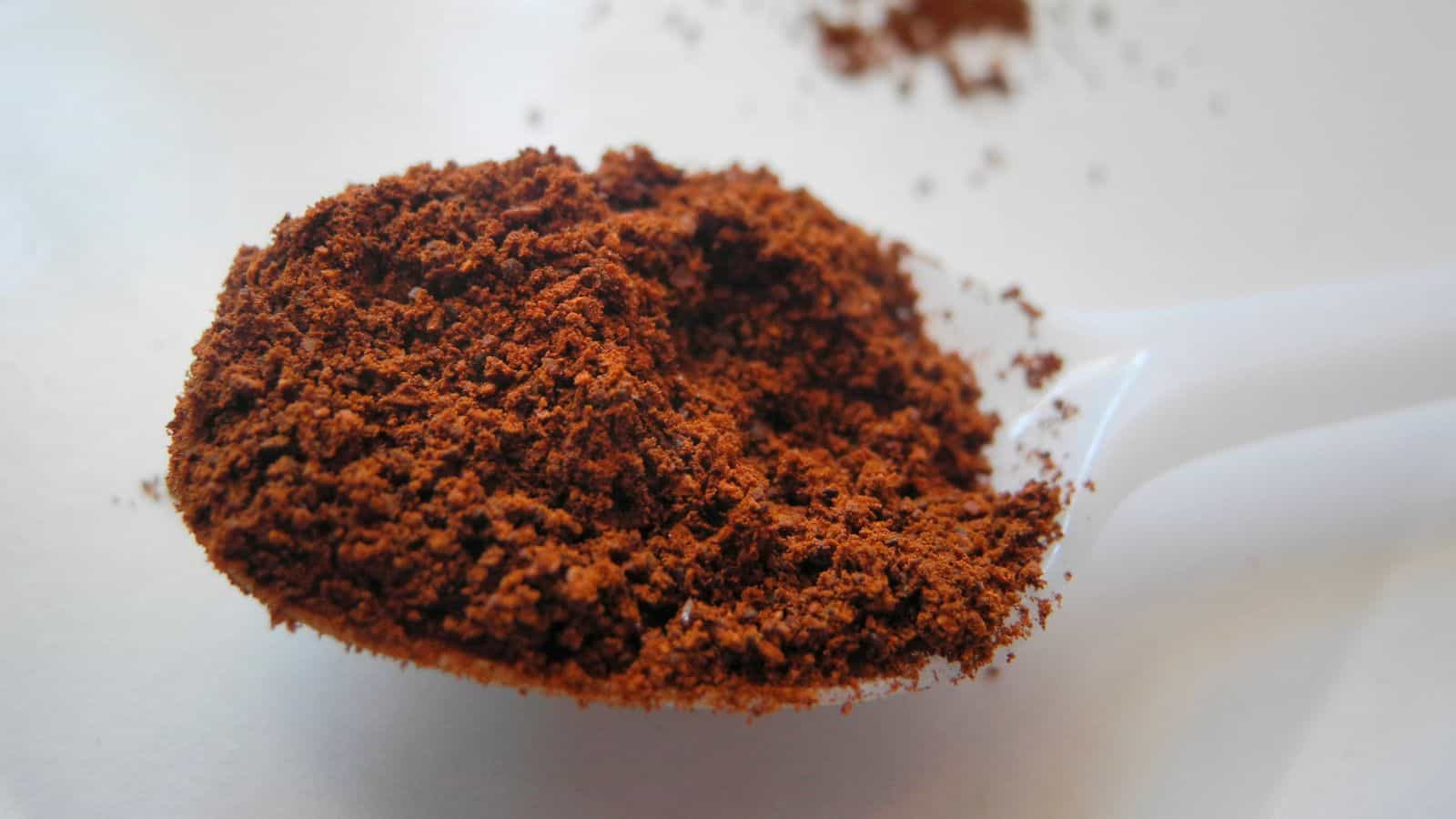 Close-up of a white plastic spoon containing brown ground coffee. Some coffee grounds are scattered on a white surface in the background.