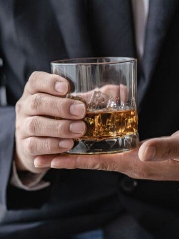 A person in a suit holds a glass of whiskey with ice cubes. The focus is on the hands and the glass, with a blurred background.