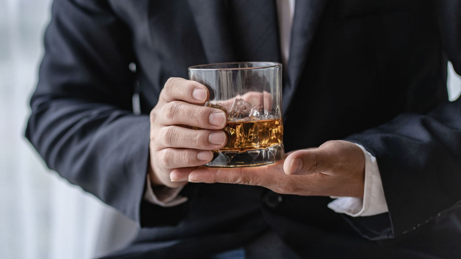 A person in a suit holds a glass of whiskey with ice cubes. The focus is on the hands and the glass, with a blurred background.