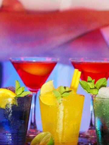 Several colorful cocktails are lined up on a bar counter. In the background, a person with crossed arms is standing, wearing a white shirt. The drinks have garnishes like mint leaves, citrus slices, and vibrant straws.