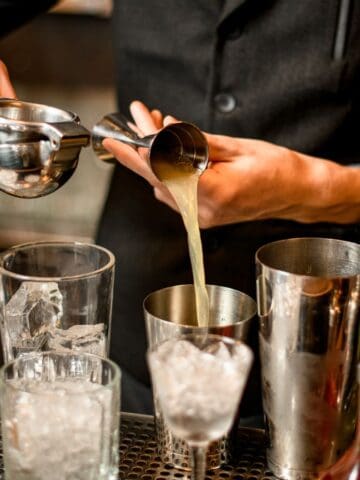 A bartender in a dark vest is squeezing juice into a jigger over a row of cocktail glasses filled with ice. Several metal shakers and bottles are arranged in front of them on the bar counter.