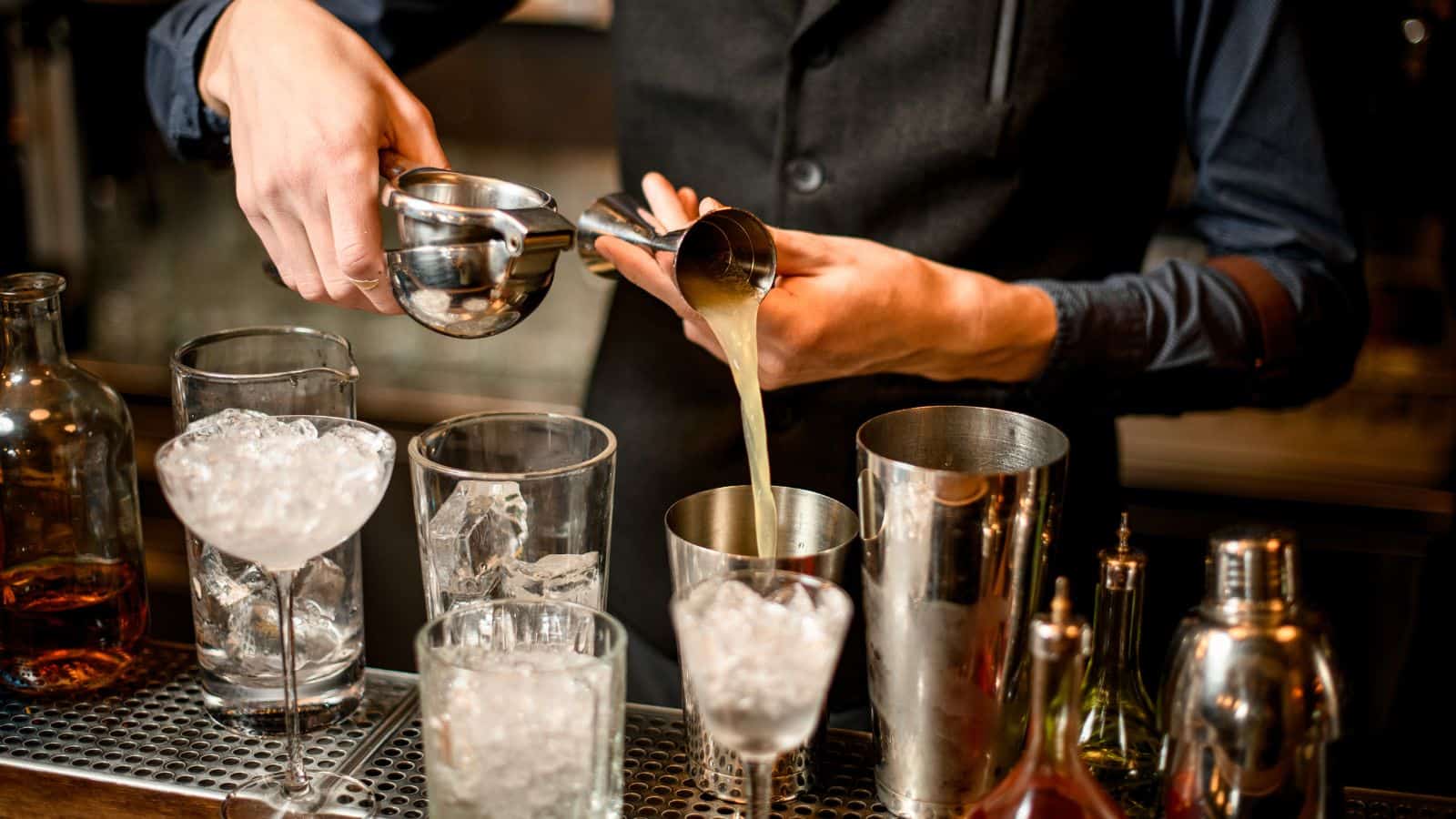 A bartender in a dark vest is squeezing juice into a jigger over a row of cocktail glasses filled with ice. Several metal shakers and bottles are arranged in front of them on the bar counter.