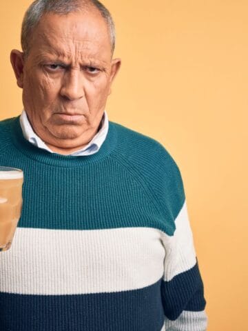 Elderly man with a serious expression holding a mug of beer. He is wearing a teal and white striped sweater, standing against a plain orange background.