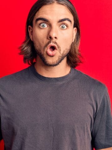 A man with long hair and a surprised expression holds a plastic cup of iced beverage with a straw. He is wearing a dark t-shirt and stands against a bright red background.