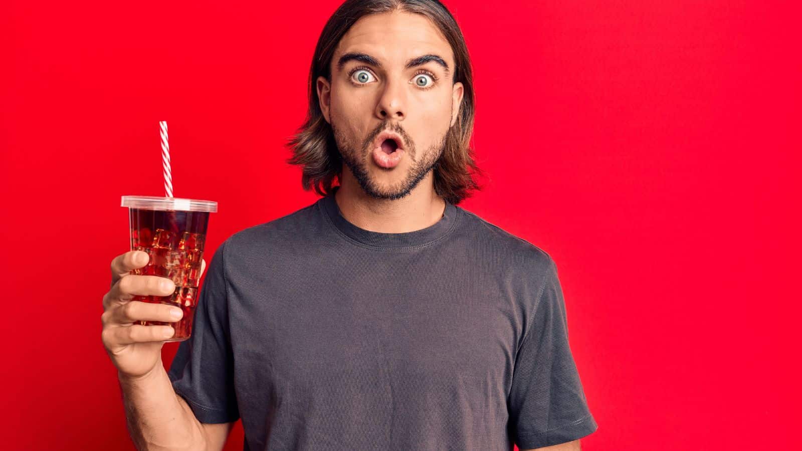 A man with long hair and a surprised expression holds a plastic cup of iced beverage with a straw. He is wearing a dark t-shirt and stands against a bright red background.