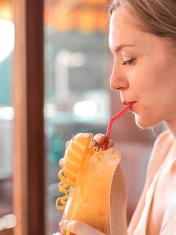 A person with light brown hair is sitting and sipping a yellow drink through a red straw. The drink is in a tall glass with a decorative lemon slice and curly straw. They are indoors, next to a window with sunlight streaming in.