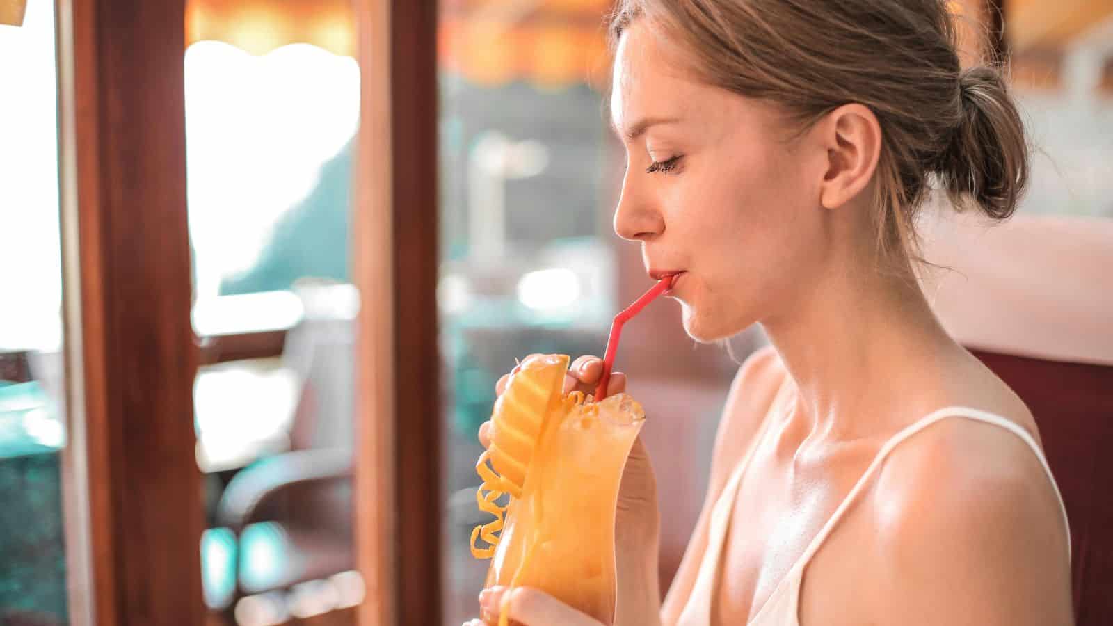 A person with light brown hair is sitting and sipping a yellow drink through a red straw. The drink is in a tall glass with a decorative lemon slice and curly straw. They are indoors, next to a window with sunlight streaming in.