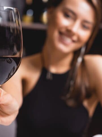 A woman in a black sleeveless top is sitting in front of wine shelves, smiling and holding a glass of red wine towards the camera. The background shows blurred bottles on the shelves.