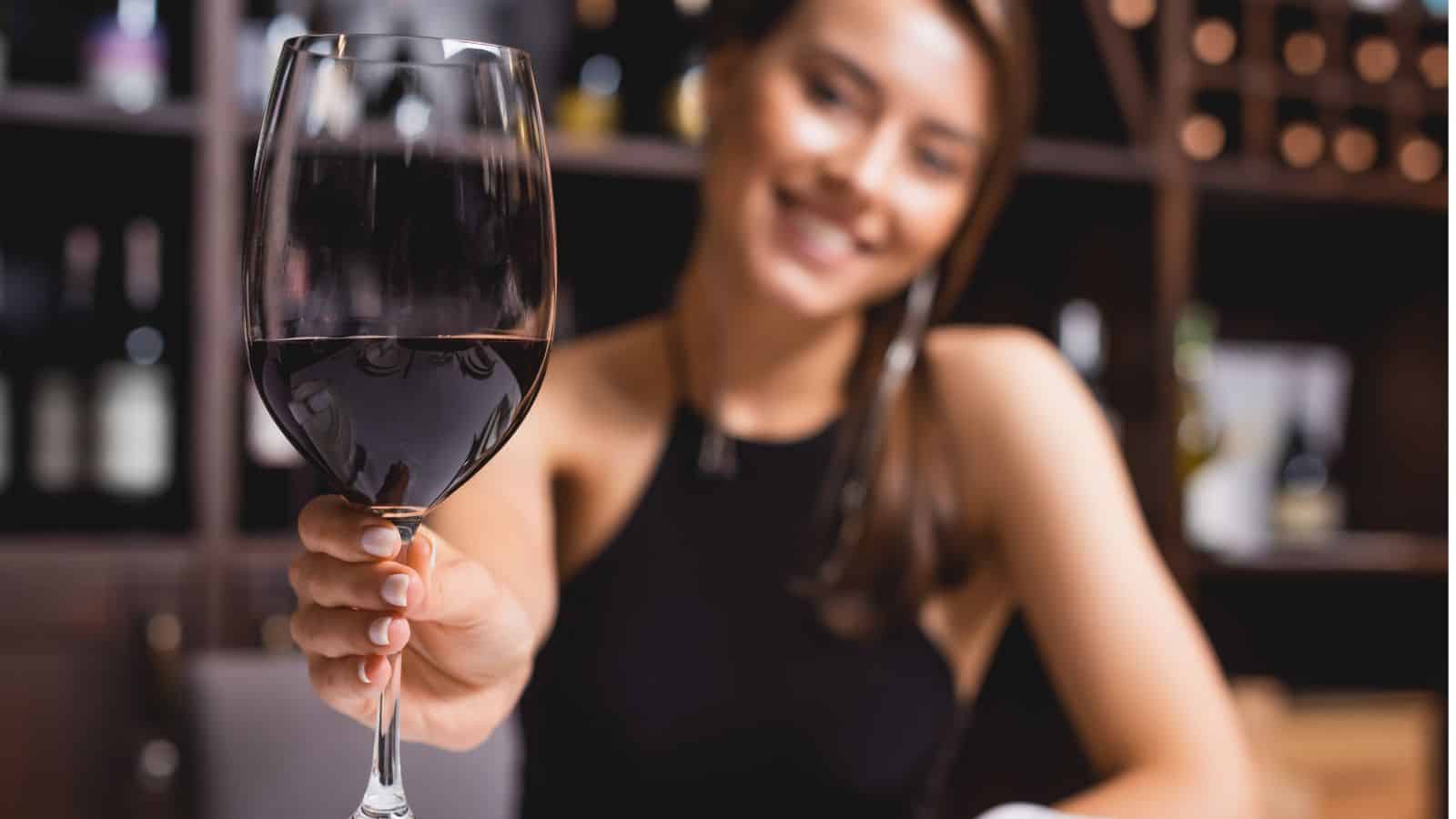 A woman in a black sleeveless top is sitting in front of wine shelves, smiling and holding a glass of red wine towards the camera. The background shows blurred bottles on the shelves.