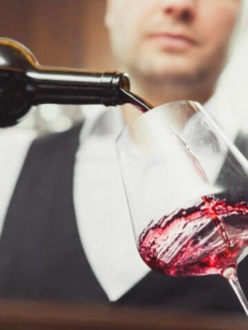 A person in a black vest and white shirt pours red wine from a bottle into a wine glass. The background is blurred, focusing on the wine being poured.