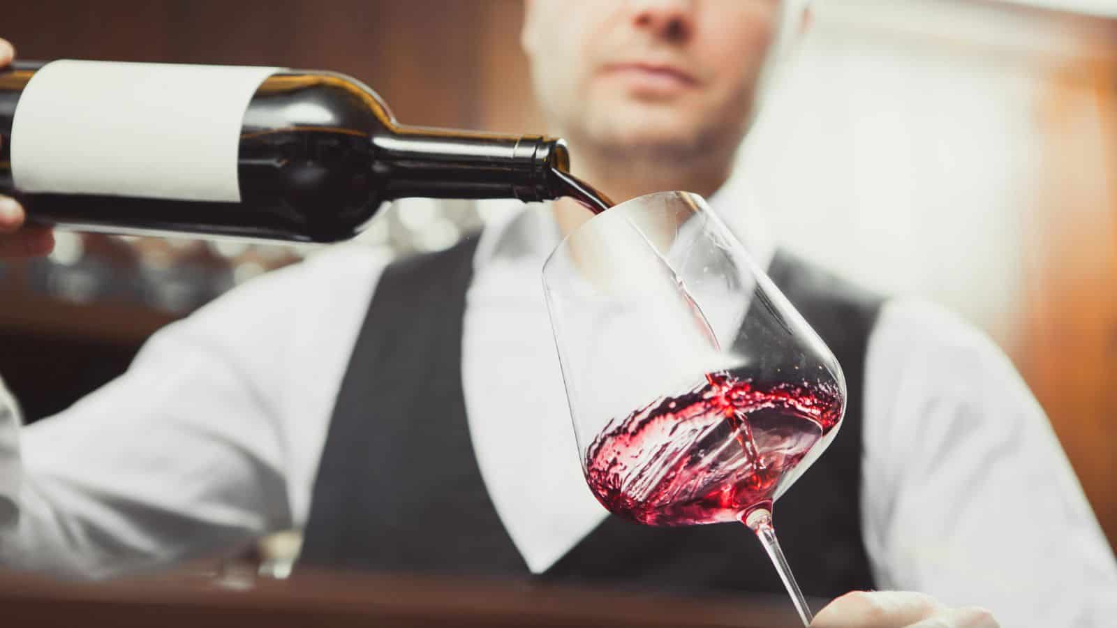 A person in a black vest and white shirt pours red wine from a bottle into a wine glass. The background is blurred, focusing on the wine being poured.