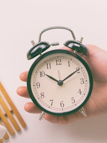 A hand holds a black and white analog alarm clock showing 10:10. Next to it is a cup filled with dark liquid, possibly coffee, placed on a wooden slatted mat. The background is a plain, light surface.