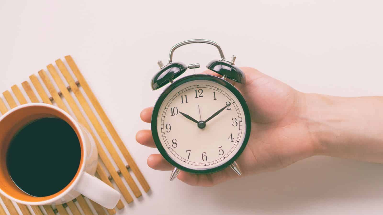 A hand holds a black and white analog alarm clock showing 10:10. Next to it is a cup filled with dark liquid, possibly coffee, placed on a wooden slatted mat. The background is a plain, light surface.