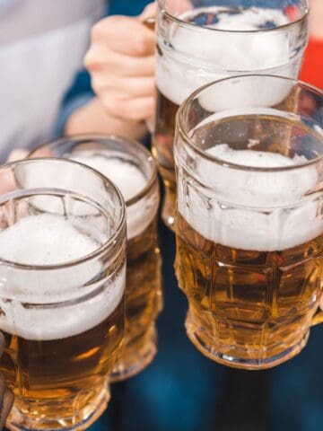 Four people clink glass mugs filled with beer, forming a toast. The mugs contain frothy beer, and the group is a mix of diverse individuals. They are wearing casual clothing, with blurred background suggesting an indoor setting.
