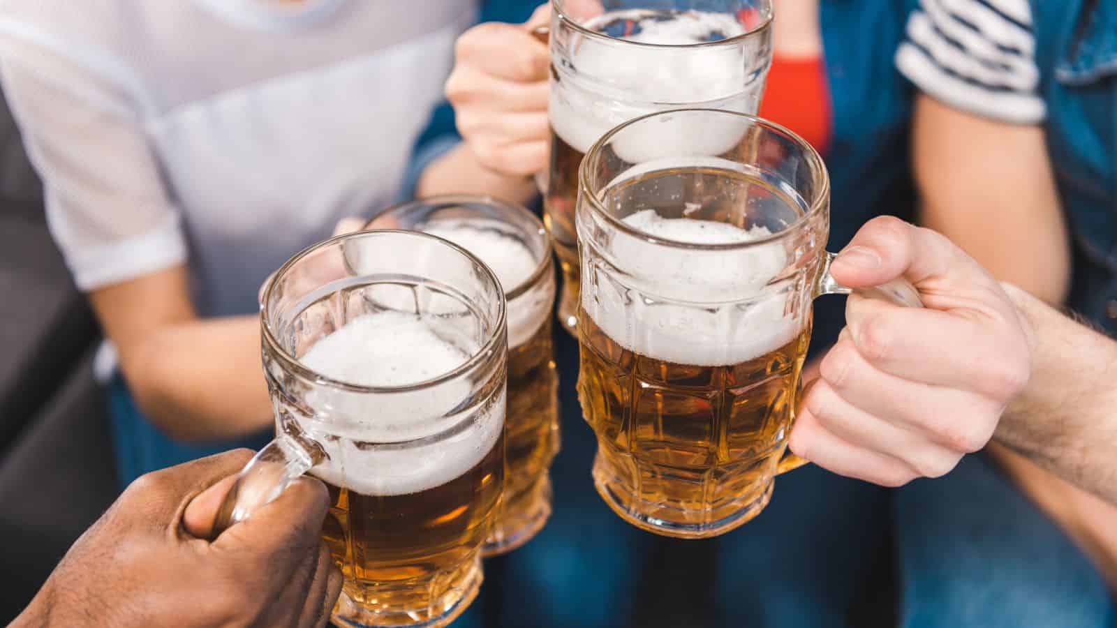 Four people clink glass mugs filled with beer, forming a toast. The mugs contain frothy beer, and the group is a mix of diverse individuals. They are wearing casual clothing, with blurred background suggesting an indoor setting.
