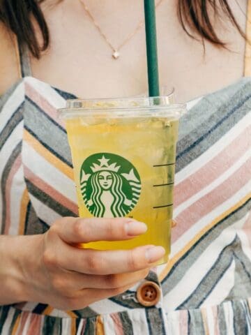 A person in a striped dress holds a clear plastic cup with a green straw. The cup contains a yellow iced beverage and has a Starbucks logo on it. The background is blurred.