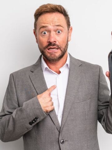 A man with a surprised expression stands against a white background. He is wearing a gray suit and white shirt. He holds a wine bottle in his right hand and points to himself with his left hand.