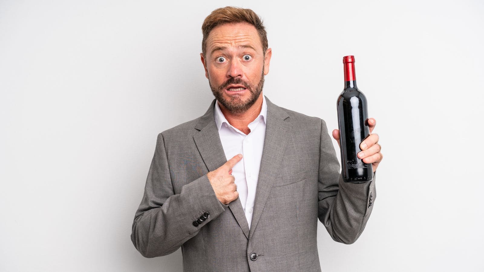 A man with a surprised expression stands against a white background. He is wearing a gray suit and white shirt. He holds a wine bottle in his right hand and points to himself with his left hand.