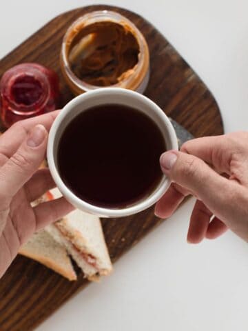 A person holds a cup of dark tea above a wooden board with sandwich triangles, a jar of peanut butter, and a jar of red jam. A glass teapot with more tea is partially visible on the right.