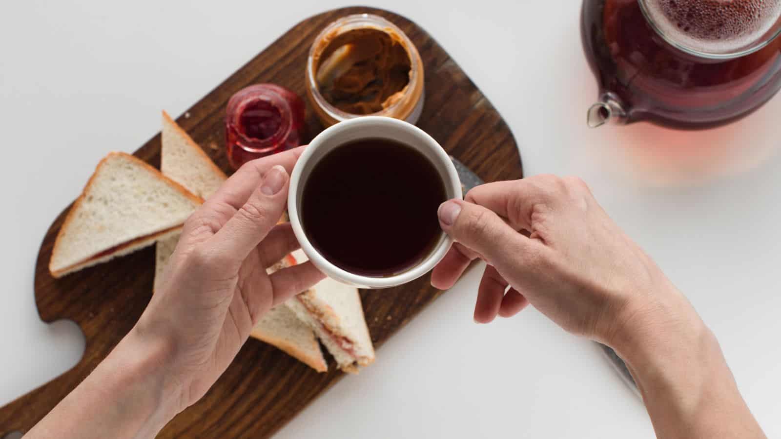 A person holds a cup of dark tea above a wooden board with sandwich triangles, a jar of peanut butter, and a jar of red jam. A glass teapot with more tea is partially visible on the right.