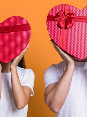 Two people wearing white shirts hold large red heart-shaped boxes in front of their faces. The boxes are decorated with ribbons. The background is orange.