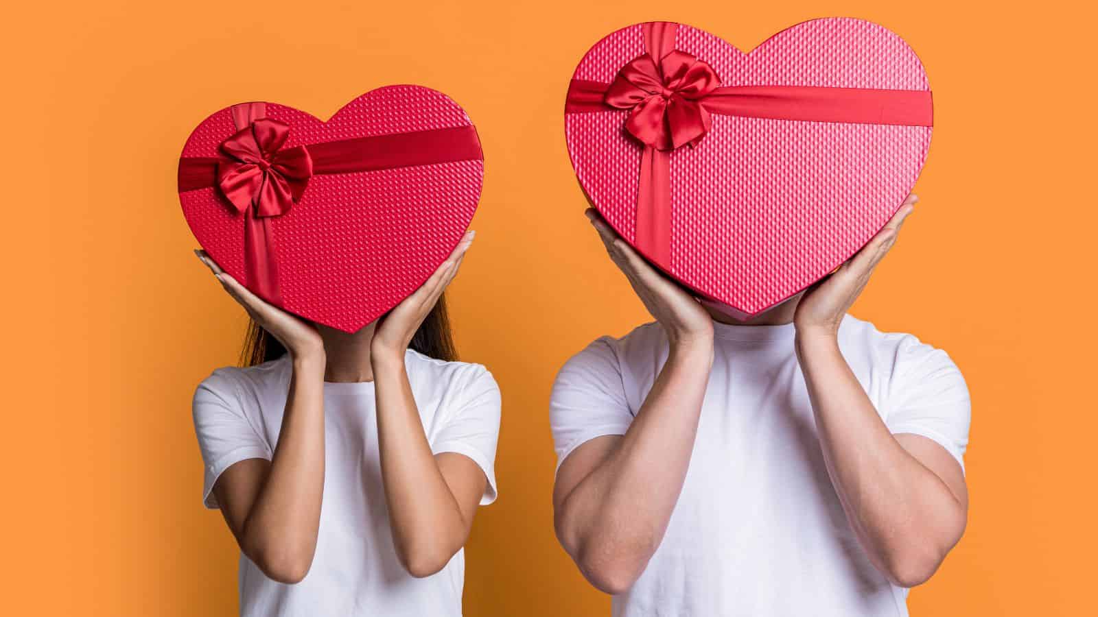 Two people wearing white shirts hold large red heart-shaped boxes in front of their faces. The boxes are decorated with ribbons. The background is orange.