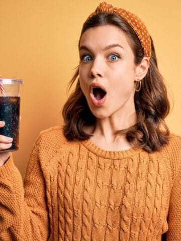A woman with an orange sweater and headband holds a large cup with a striped straw, looking surprised. The background is a solid yellow color.