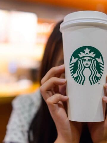 A person holds a large Starbucks coffee cup in front of their face. The background is blurred, showing warm-toned decor typical of a coffee shop setting.