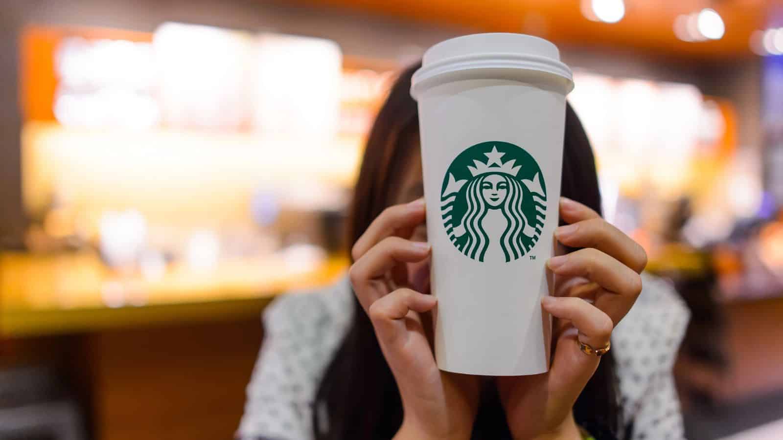 A person holds a large Starbucks coffee cup in front of their face. The background is blurred, showing warm-toned decor typical of a coffee shop setting.