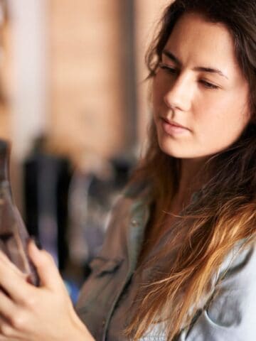 A woman with long brown hair is standing in a store aisle, looking at a packaged product. She is wearing a light denim shirt and appears focused on the item in her hand. Shelves filled with similar products are visible around her.