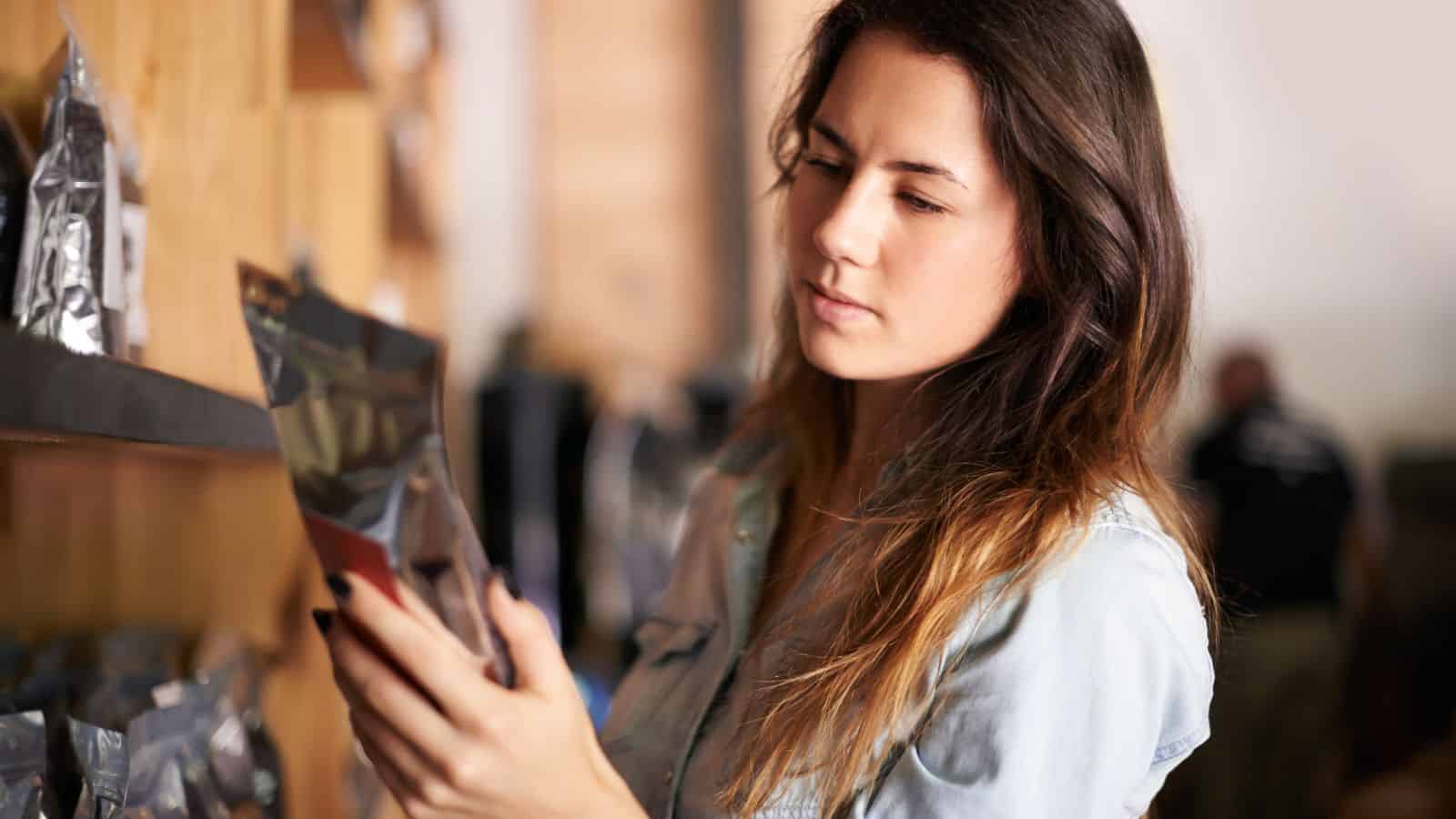 A woman with long brown hair is standing in a store aisle, looking at a packaged product. She is wearing a light denim shirt and appears focused on the item in her hand. Shelves filled with similar products are visible around her.