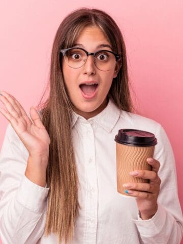 A surprised person with long brown hair and glasses is holding a paper coffee cup. They are wearing a white button-up shirt and standing against a pink background, with their mouth open and one hand raised.