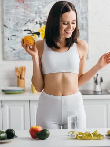 A woman in a kitchen, wearing white workout clothes, stands at a counter holding an orange. There are various fruits and vegetables, a blender, and a measuring tape on the counter. A colorful abstract painting hangs on the wall behind her.