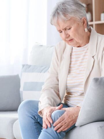 An elderly woman with short gray hair sits on a light-colored sofa with a pained expression. She is wearing a light blazer and striped shirt and is holding her knee with both hands, indicating discomfort. The room is bright with shelves in the background.