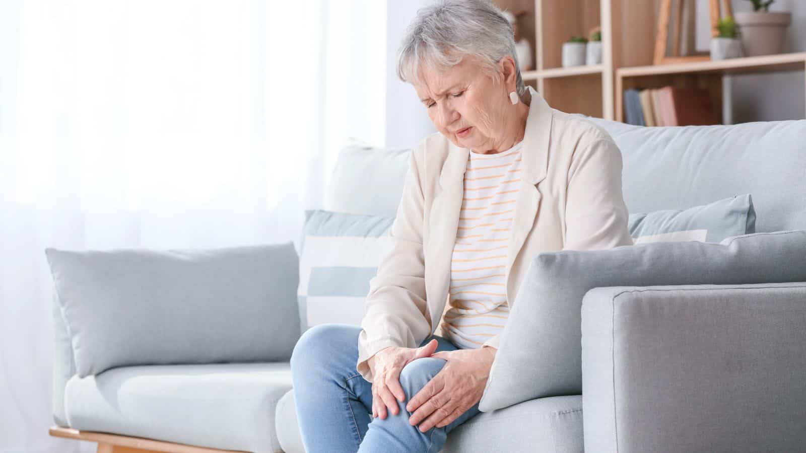 An elderly woman with short gray hair sits on a light-colored sofa with a pained expression. She is wearing a light blazer and striped shirt and is holding her knee with both hands, indicating discomfort. The room is bright with shelves in the background.