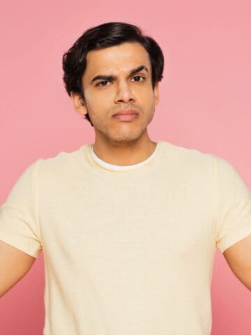 A man in a yellow shirt stands against a pink background, holding a yellow cup with a red lid in one hand and a brown cup with a white lid in the other, looking confused.