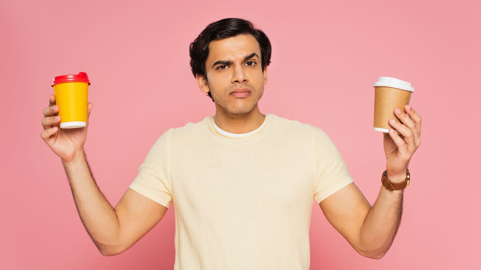 A man in a yellow shirt stands against a pink background, holding a yellow cup with a red lid in one hand and a brown cup with a white lid in the other, looking confused.