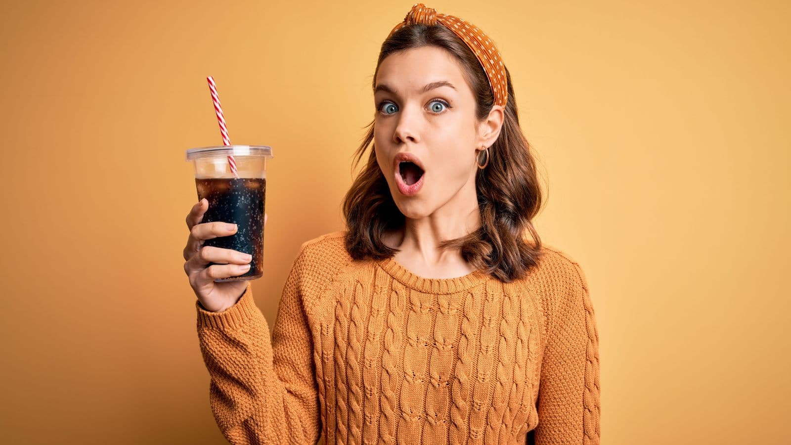 A woman with an orange sweater and headband holds a large cup with a striped straw, looking surprised. The background is a solid yellow color.