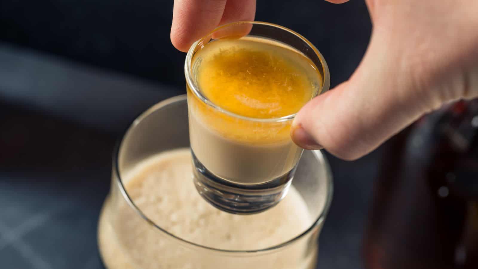 A hand holds a shot glass filled with a yellow and cream-colored layered beverage above a partially visible glass of creamy liquid. The background is blurred, with dark tones.