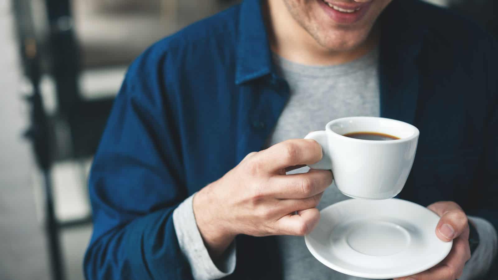 A person in a blue jacket and gray shirt holds a white cup of coffee in one hand and a saucer in the other. The background is blurred, and the person is smiling slightly.