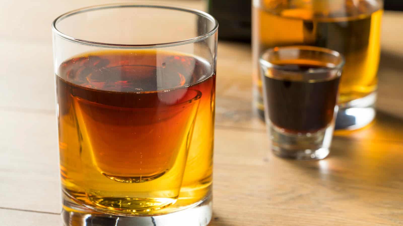 Three glasses containing amber or dark brown liquids sit on a wooden surface. The foreground features a large glass mostly filled, while the background includes a smaller shot glass and another partially visible glass.