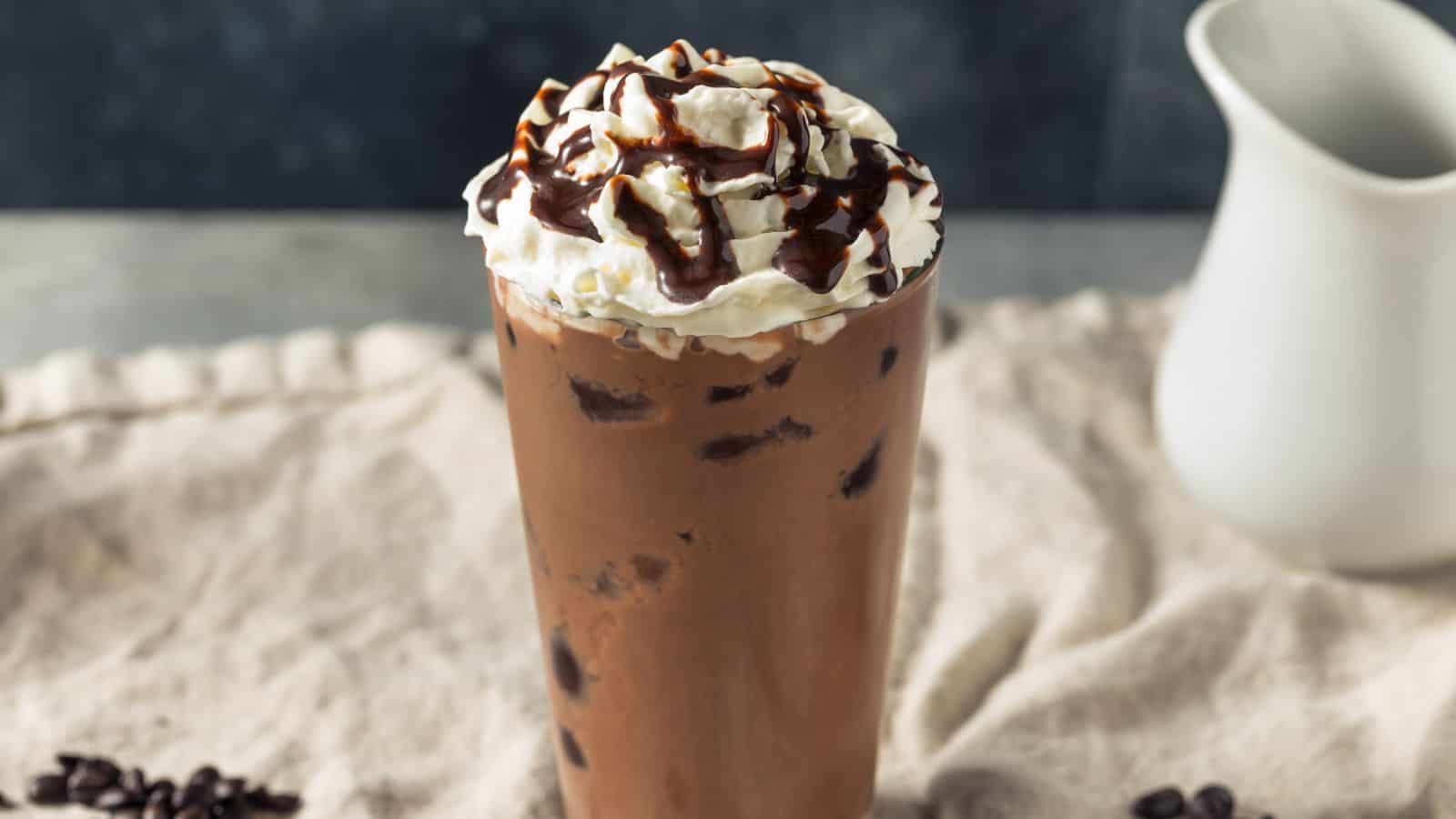 A tall glass of iced mocha topped with whipped cream and drizzled with chocolate syrup sits on a beige cloth. A few coffee beans are scattered nearby, and a white pitcher is in the background.