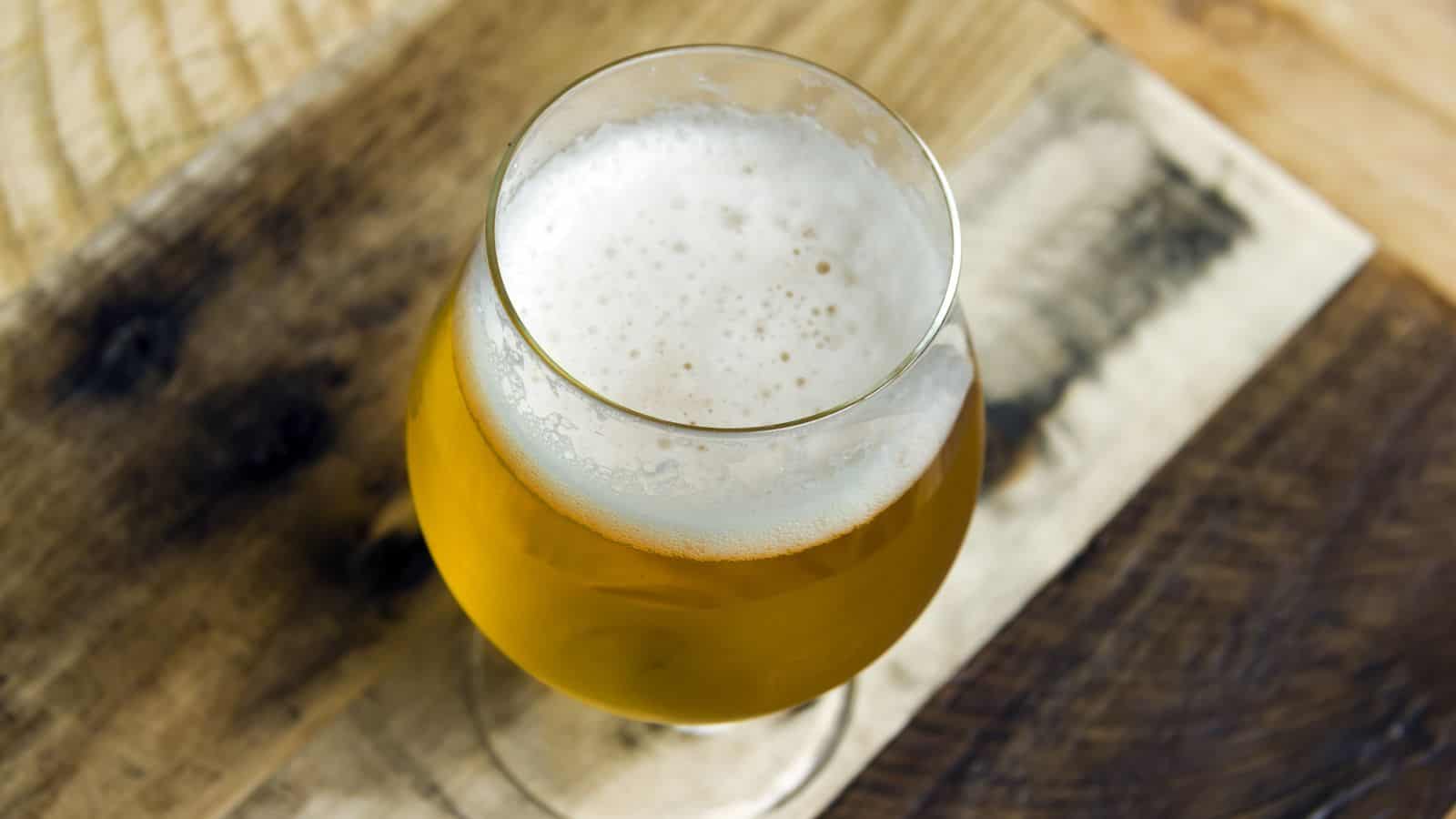 A glass of amber-colored beer with a frothy head sits on a rustic wooden table, viewed from above.