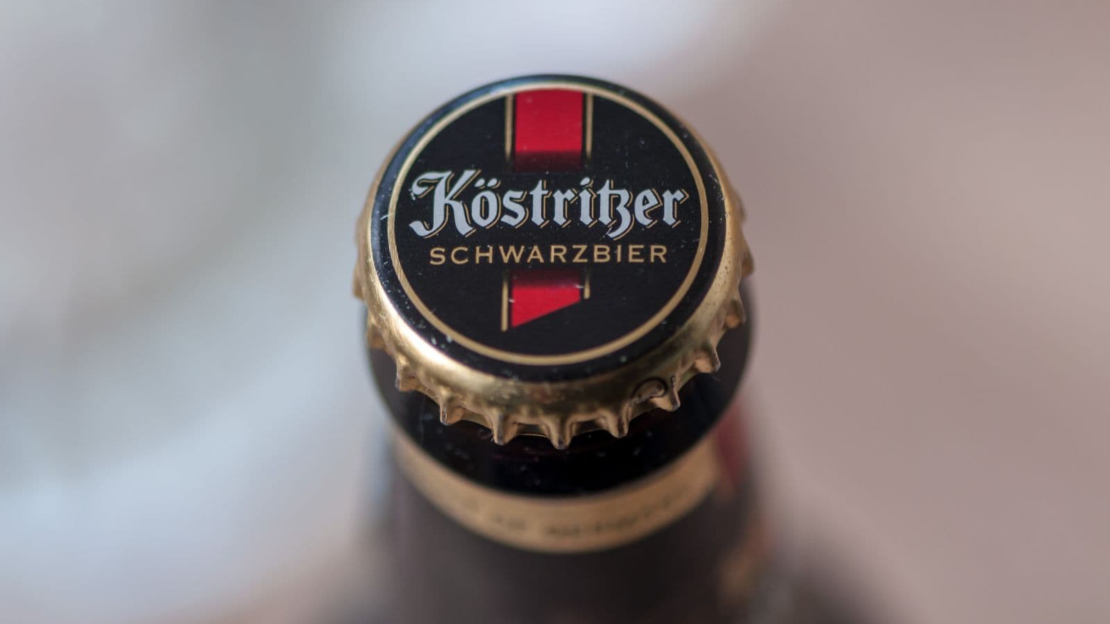 Close-up of a K&ouml;stritzer Schwarzbier bottle cap. The cap is black with the K&ouml;stritzer logo and red banners. The text "Schwarzbier" is visible beneath the logo. The cap is attached to the top of a dark bottle.