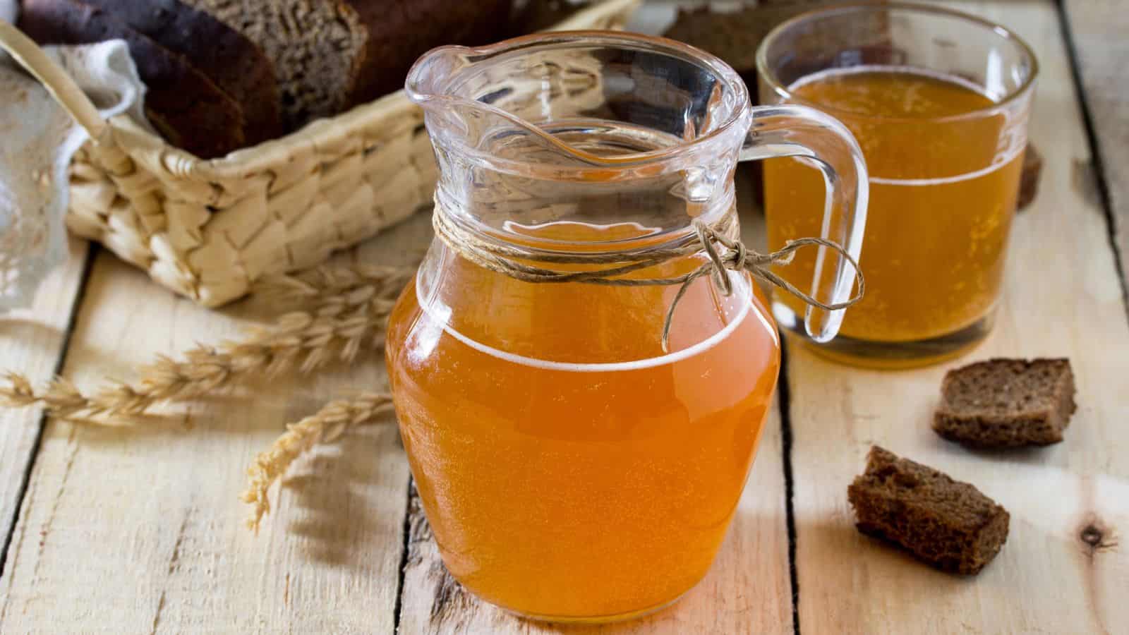 A glass pitcher filled with a light amber liquid sits on a wooden table. Next to it is a glass with the same liquid. Nearby are pieces of dark bread and a basket with a loaf of brown bread. Wheat stalks are placed beside the pitcher.