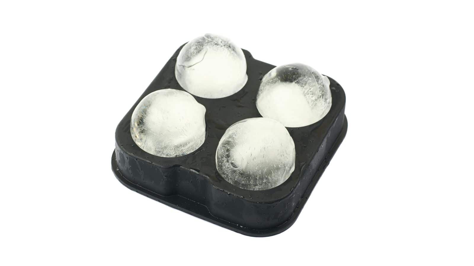 A black ice mold tray containing four round ice spheres is positioned against a plain white background. The ice balls appear clear and solid, sitting snugly within the tray.